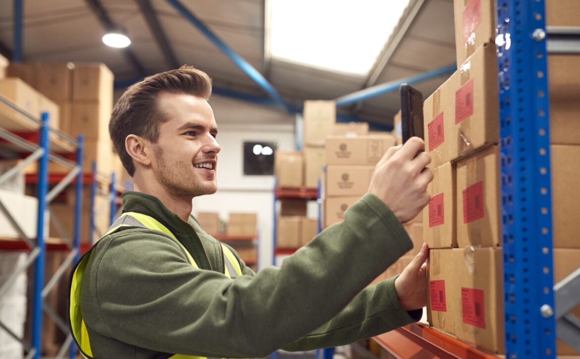 Male Worker Wearing Inside Warehouse Scanning Stock Barcode On Shelves Using Digital Device Or Phone